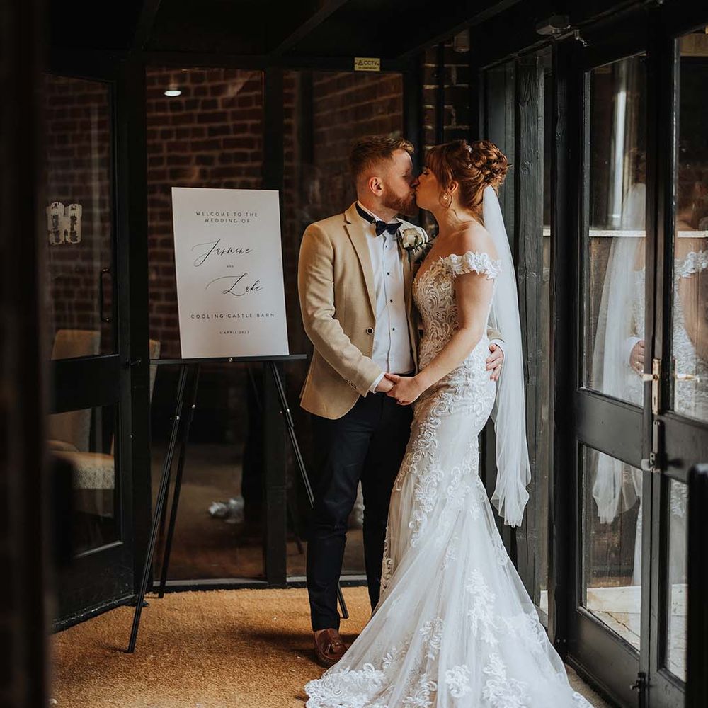 Bride and groom kiss next to black and white welcome wedding sign with personalised details