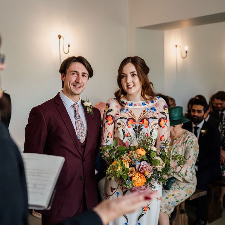 Groom in three piece burgundy suit with blue suit and floral tie with bride in bright and colourful floral wedding dress