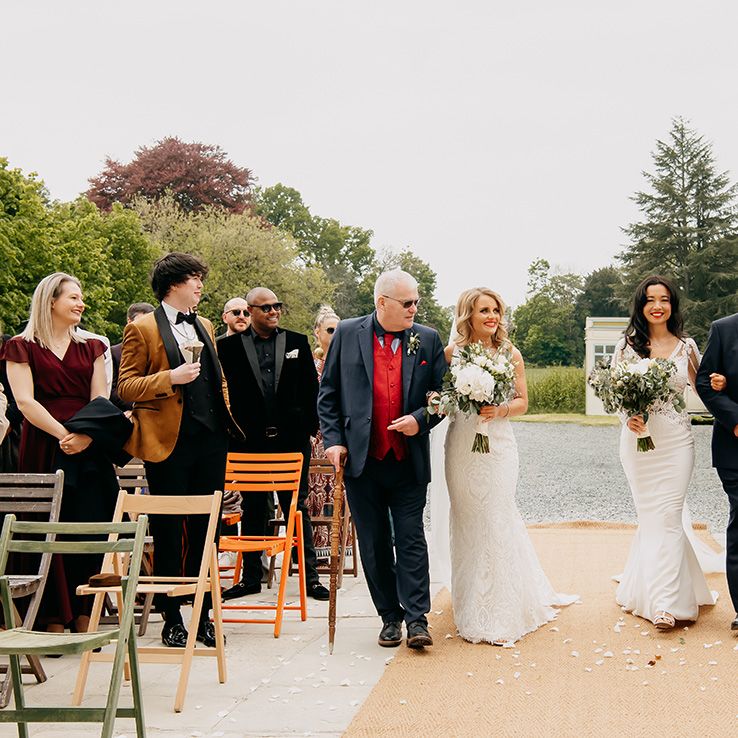 Father of the brides walk the brides down the aisle decorated with white flower petals at the Chateau De la Motte Husson