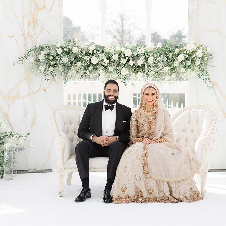 Bride wears traditional embellished saree on her wedding day as she sits beside groom wearing black tie in front of white florals 