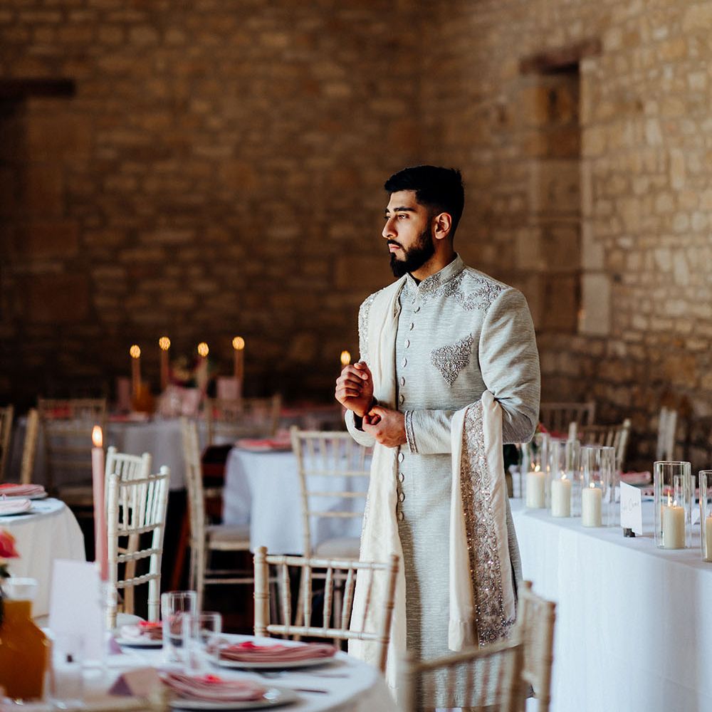 Groom wearing traditional sherwani in light grey with embellishment stands within barn for wedding reception 