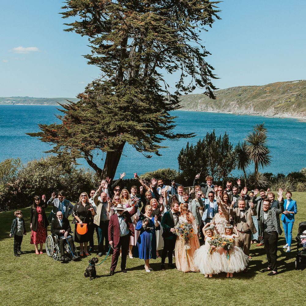 Bride & groom stand with their wedding party in front of the sea front in Cornwall for sunny wedding day outdoors