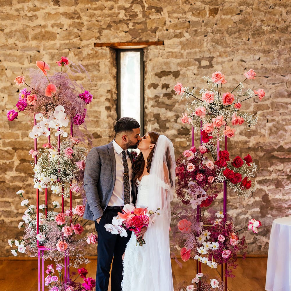 Bride & groom kiss between bright pink floral archway on their wedding day at Priston Mill