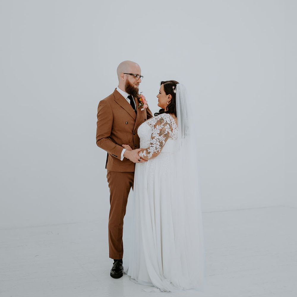 Bride wears lace embellished sheer sleeves and stands beside her groom in retro styled brown suit