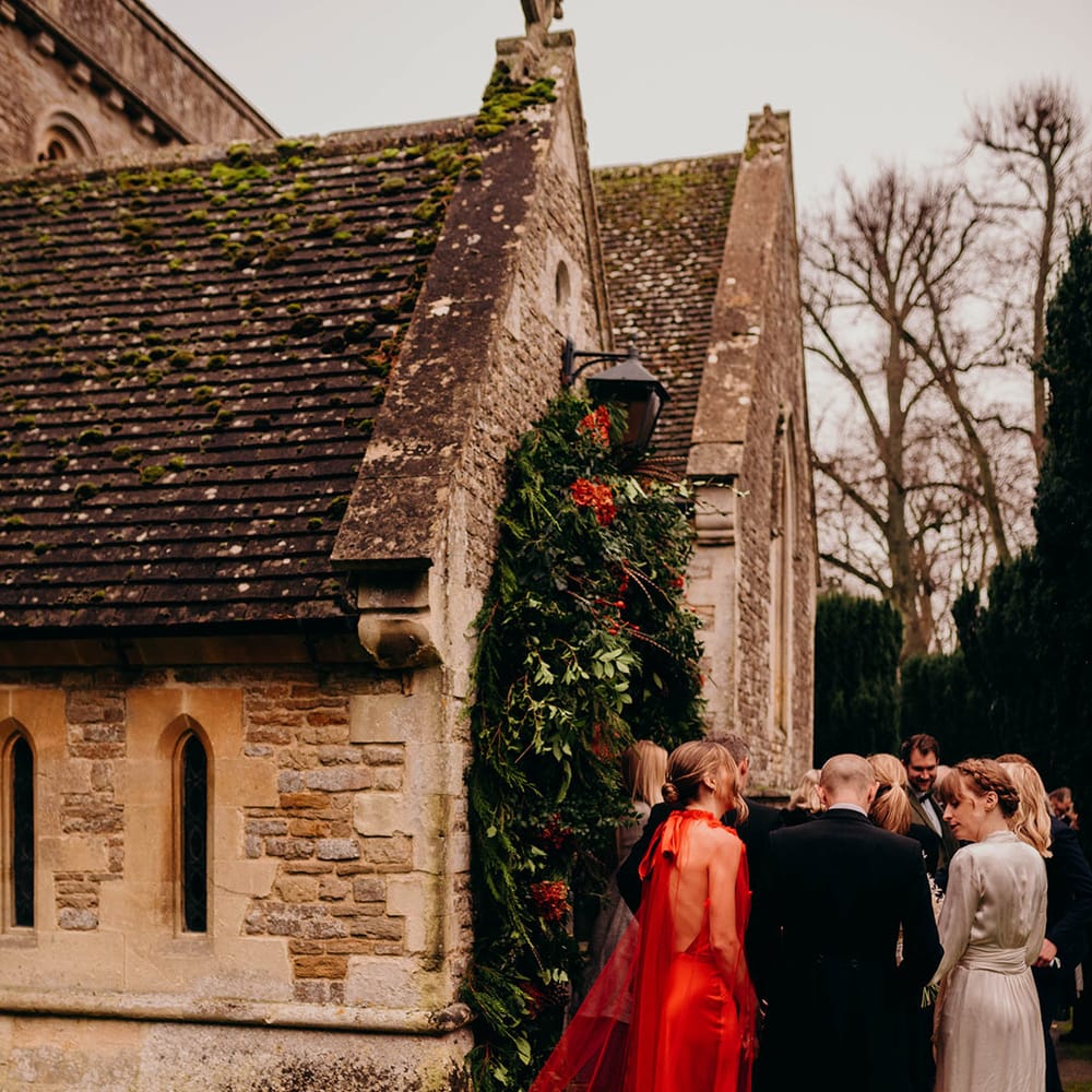 bride in satin red wedding dress with cape and other wedding guests standing by the church with large winter themed foliage and floral wedding arch