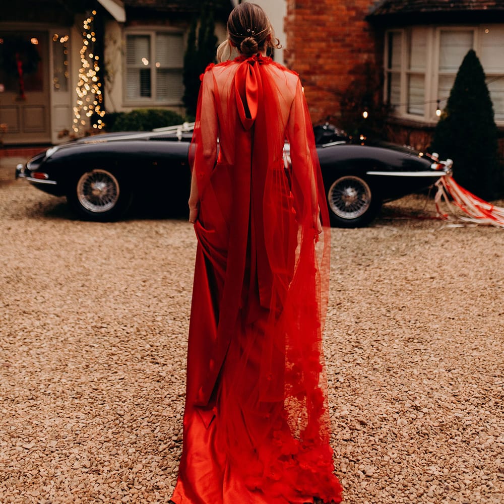 Bride in red silk wedding dress with cape, large red embellished head band and large red satin bow in front of classic convertible wedding car