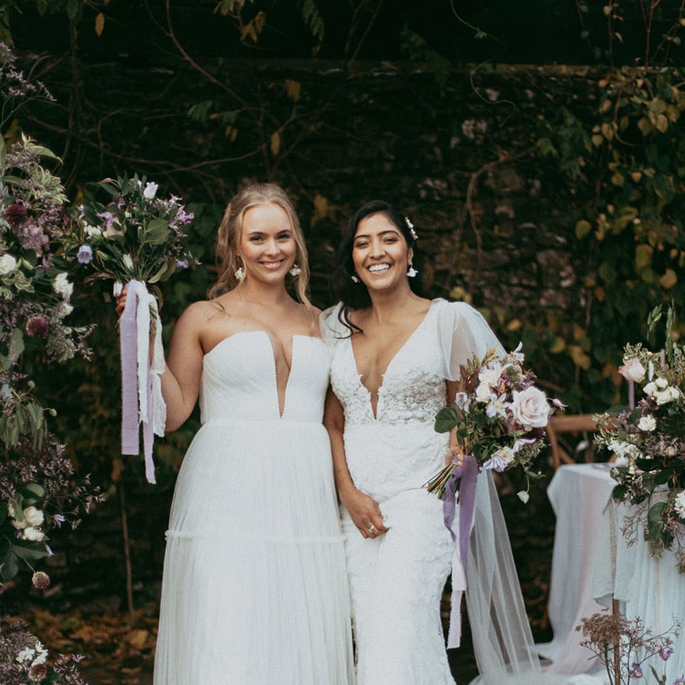 Brides in complimenting wedding dresses and Dr Martens boots smiling and laughing holding mixed floral bouquets with white carnations, purple tulips, roses and foliage 