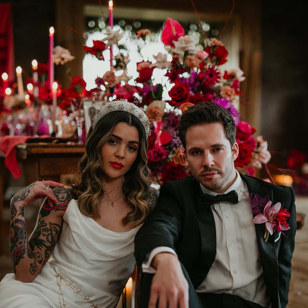 Bride in cowl neck sleeveless satin wedding dress with leg slit, white and gold glittery wedding headband, and gold and pearl body chain sitting on the floor with groom in classic black tuxedo with magenta amaryllis and orchid boutonniere surrounded by magenta florals 