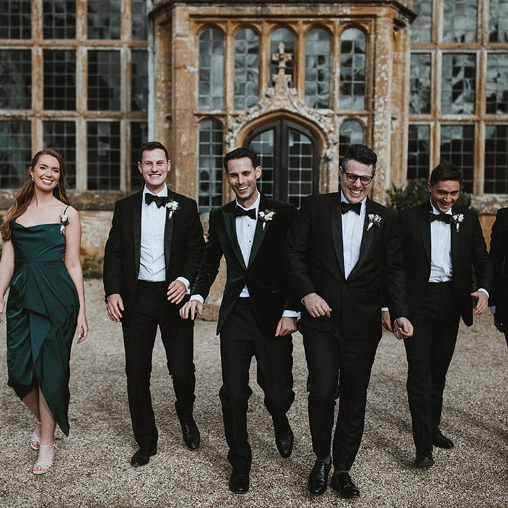 Groom and groomsmen in traditional black tuxedos with a groomswoman in a dark green cowl neck bridesmaid dress 