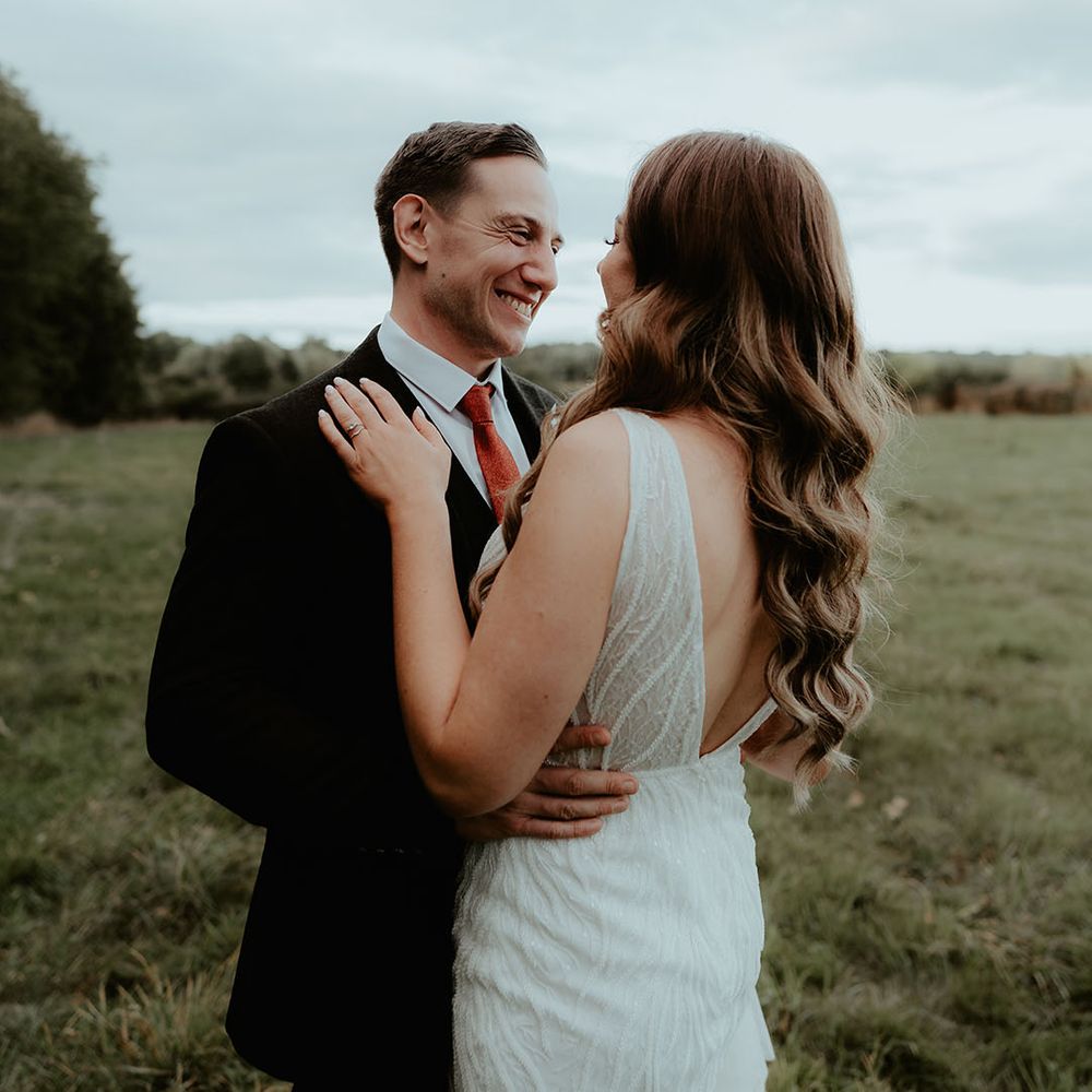 Groom smiles widely wearing a black suit and orange tie embracing the bride in a sequin wedding dress with curled hair 