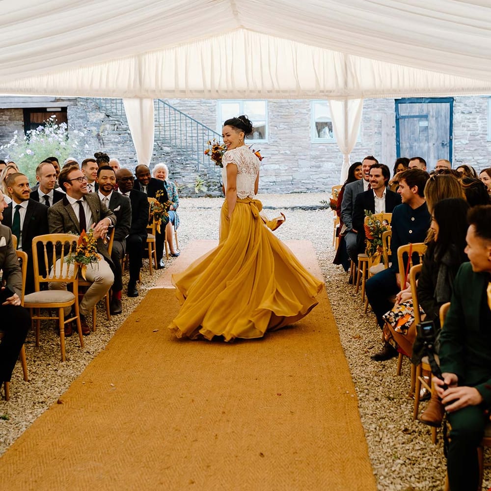 Bride in white mesh lace bridal crop top and yellow wedding skirt holding dried autumnal wedding bouquet with sunflowers, garden roses, pampas grass, eucalyptus, yellow tall kangaroo paws, orange osmanthus flowers, foliage and dried flowers walking down the aisle at Broadfield Court