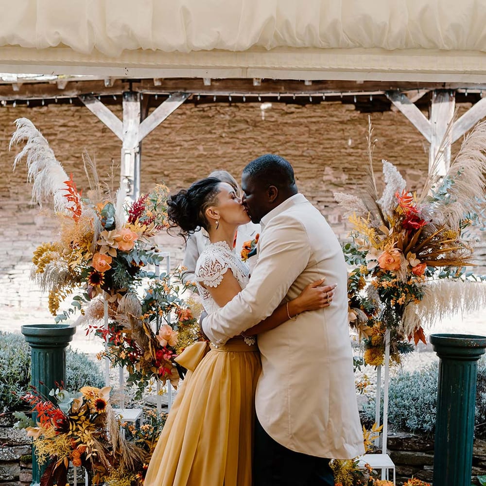 Groom in cream morning suit with forest green grooms waistcoat and trousers, forest green pocket square and orange boutonniere kissing bride in white mesh lace bridal crop top and yellow wedding skirt at the alter of Broadfield Court