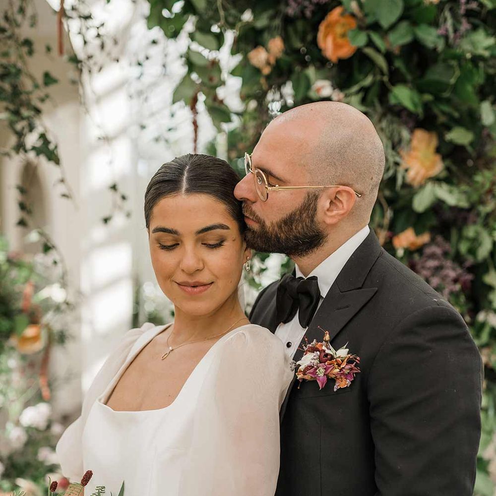 Bride in short sheer puff sleeve wedding dress with square neck and wrist loop with sheer pearl embellished bridal veil holding dried wildflower bouquet standing with groom in classic black tuxedo with bowtie and wildflower boutonniere at Buxted Park