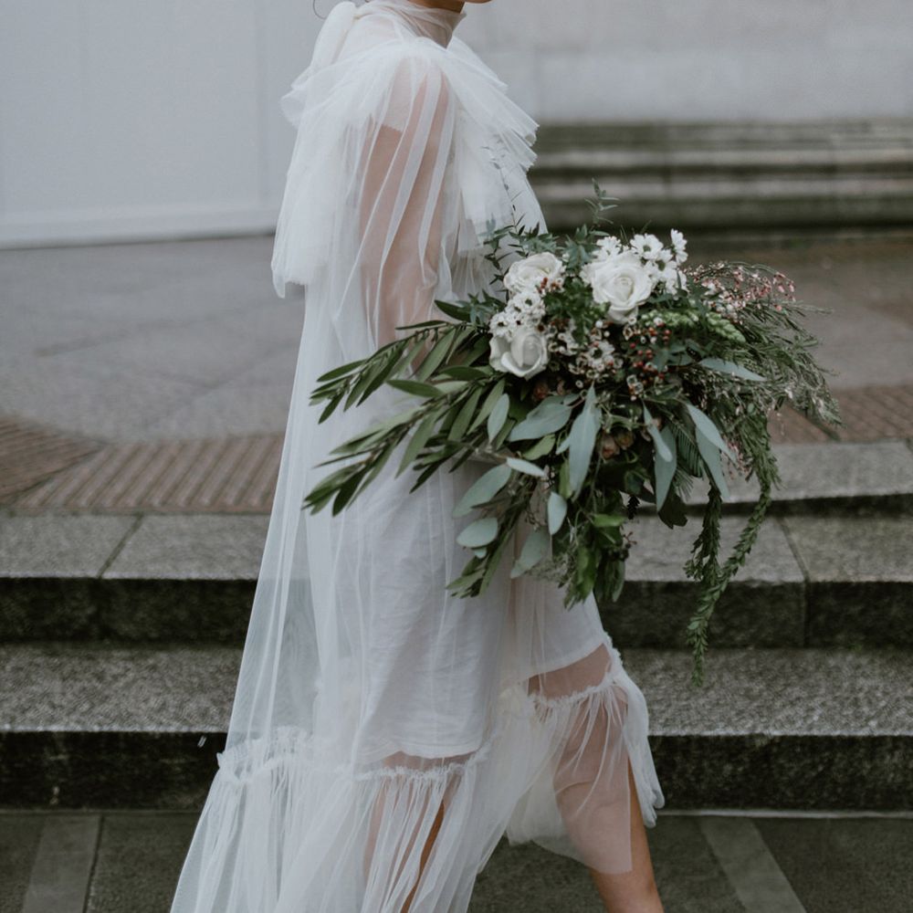 Stylish bride with short fringe and red lipstick holding a white and green bouquet whilst wearing a tulle midi dress
