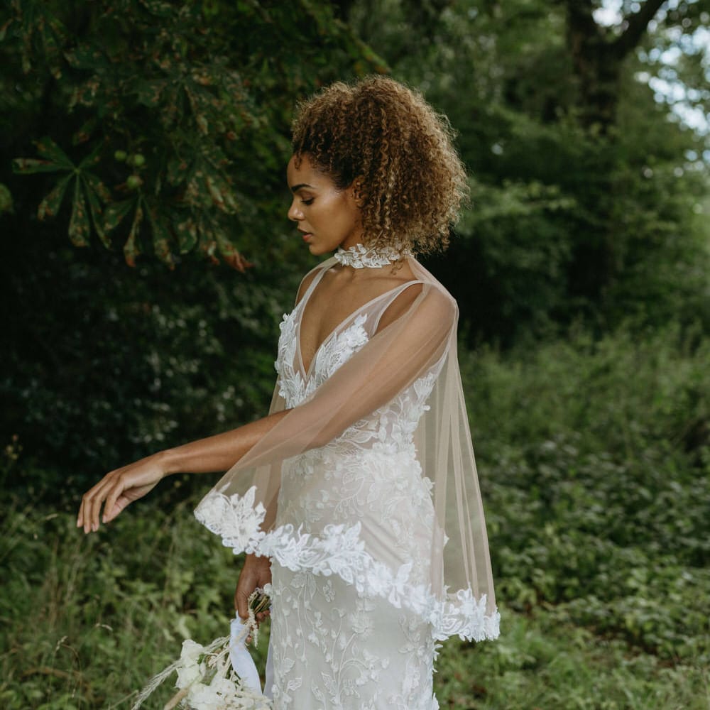 Bride wearing a floral embroidered wedding dress with matching tulle bridal cape in the woods for boho wedding at Berwick Lodge