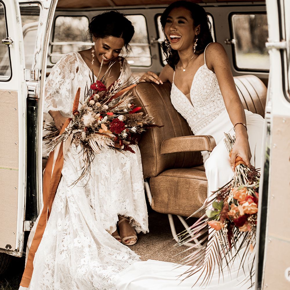 Two brides sitting in a VW camper van holding red dried and fresh flower bouquets 