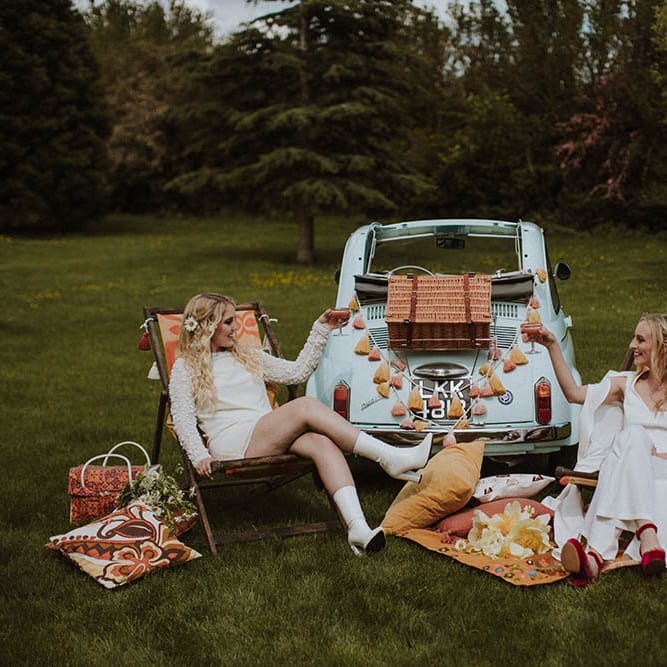 Sixties wedding inspiration with two brides sitting on deck chairs enjoying a picnic by their fiat 500