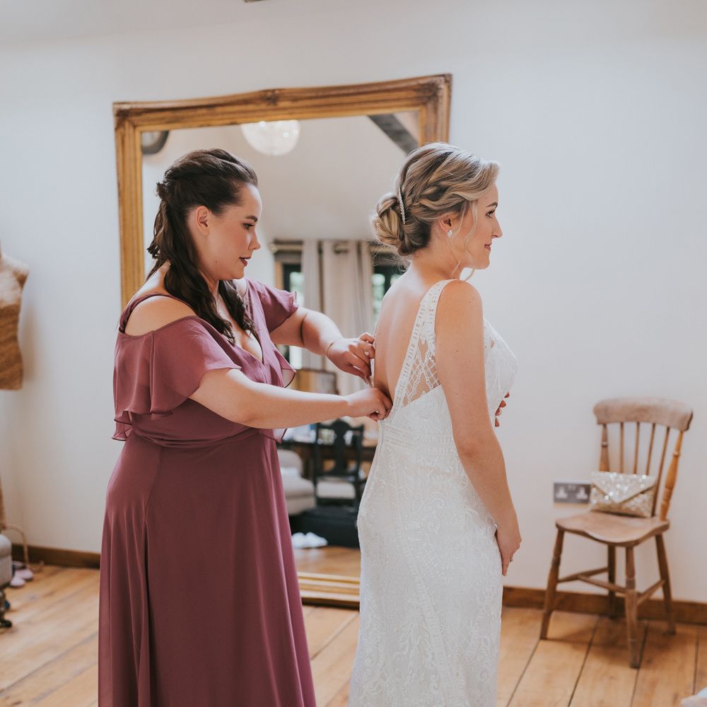 Bride in lace Justin Alexander wedding dress with train and low back gets buttoned up by bridesmaid in dusky pink bridesmaid dress before Tythe Barn wedding with barn wedding flowers