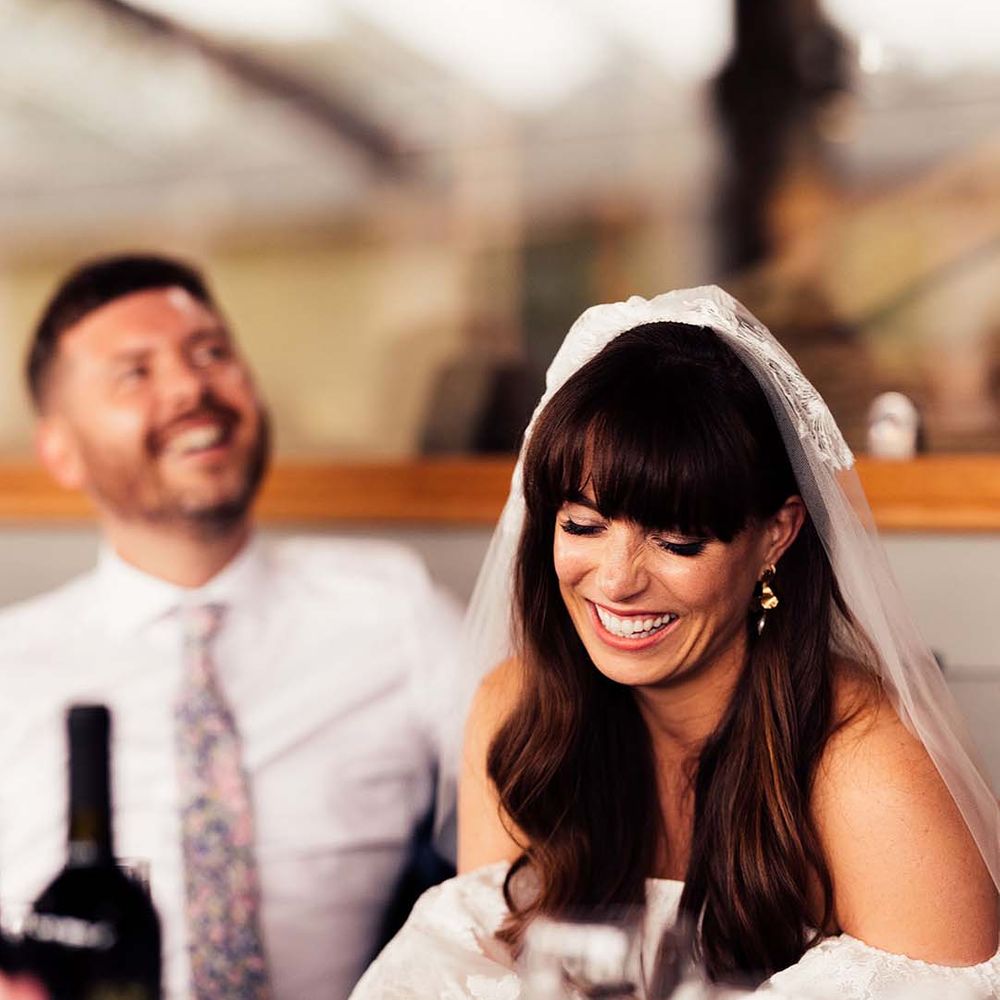 Beautiful bride with a fringe and long wavy hair wearing a veil laughing during the wedding speeches 
