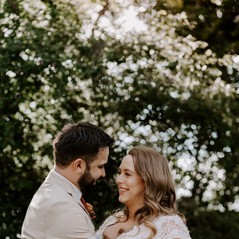 Bride in homemade lace wedding dress holding mixed rose bridal bouquet with curled hair smiles at groom in beige suit with floral buttonhole at DIY garden wedding ceremony