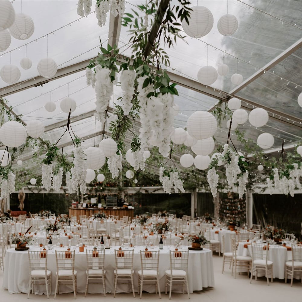 Interior of clear roofed marquee in garden with white paper lanterns and garlands hanging from the ceiling over white tables and chairs with plant decor for home garden wedding
