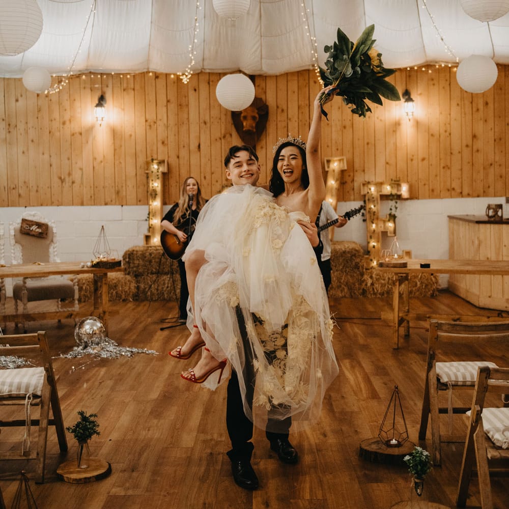 Groom in dark suit holding bride in strapless tulle wedding dress holding up bouquet for rustic wedding reception with fairy lights