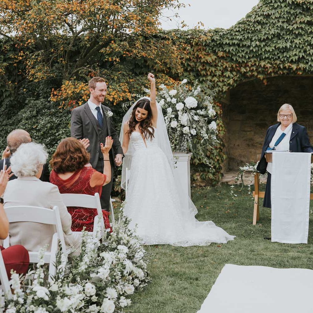 Bride in applique Pronovias wedding dress and veil throws hand in the air as she stands holding hands with groom in grey three piece suit during outdoor wedding ceremony in Buckinghamshire