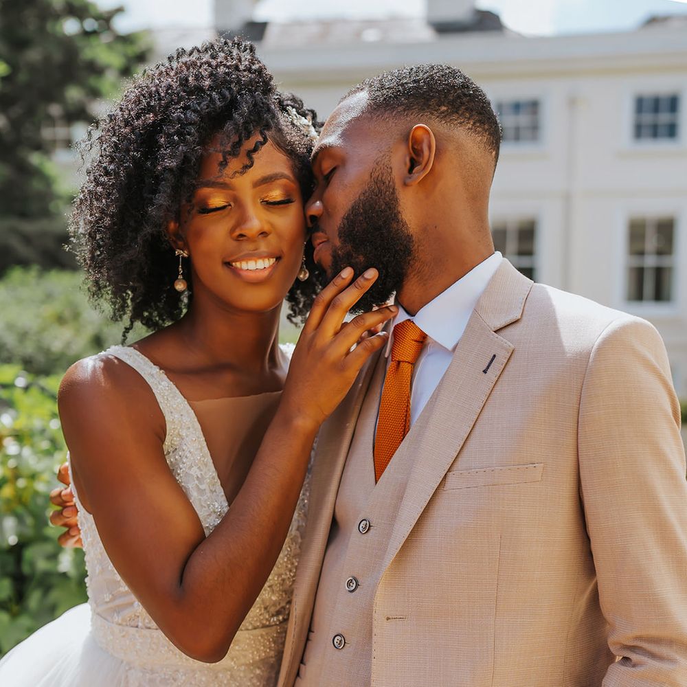 Black groom in a beige three-piece suit kissing his bride's cheek with naturally curly hair in a princess wedding dress