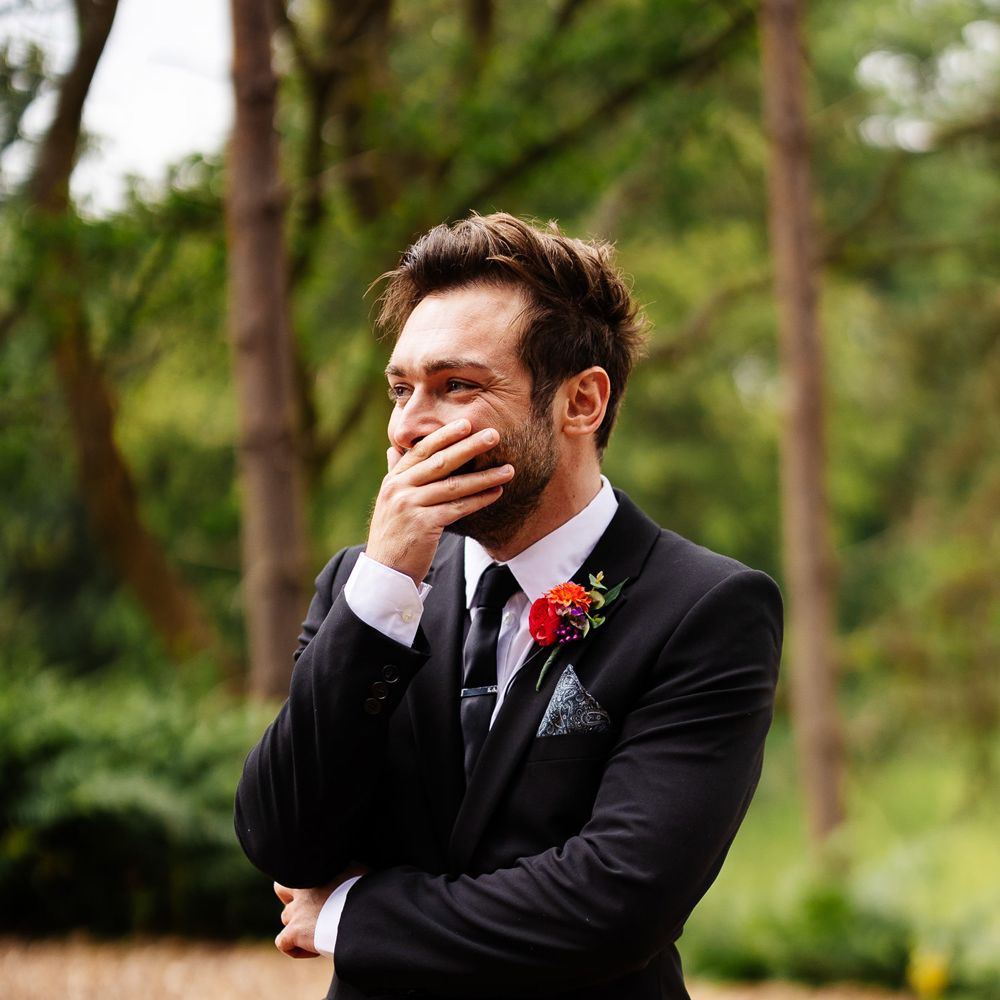 Groom in black suit and colourful floral buttonhole holds hand over his mouth as he smiles at bride walking down the aisle during outdoor woodland wedding ceremony