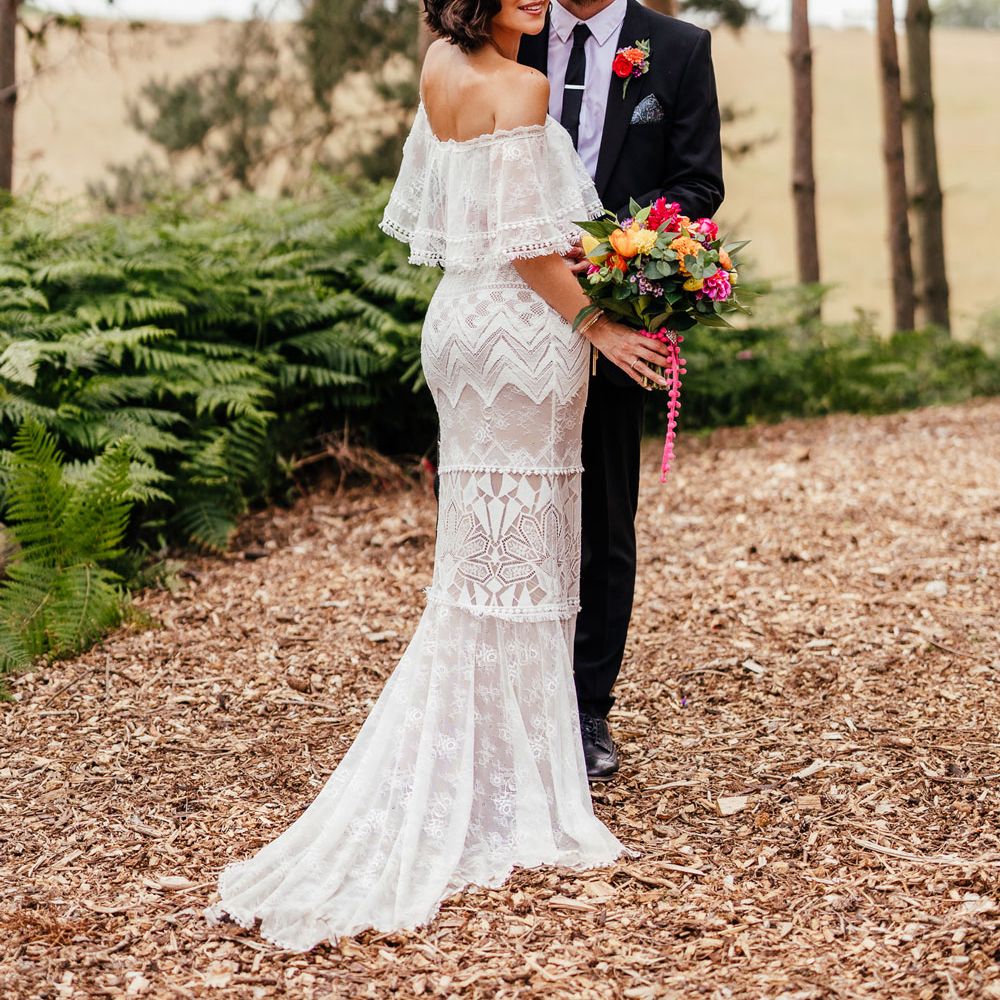Bride in lace off the shoulder Grace Loves Lace wedding dress and flower crown holds colourful wedding bouquet as she stands smiling with groom in black suit in woodland