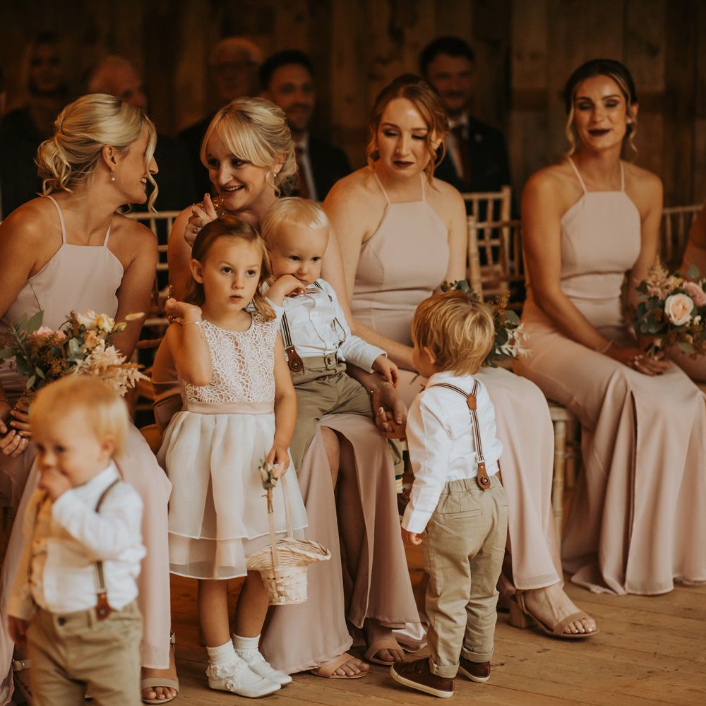 Bridesmaids in light pink halter neck bridesmaid dresses stand with flower girl and boys in white shirts and braces