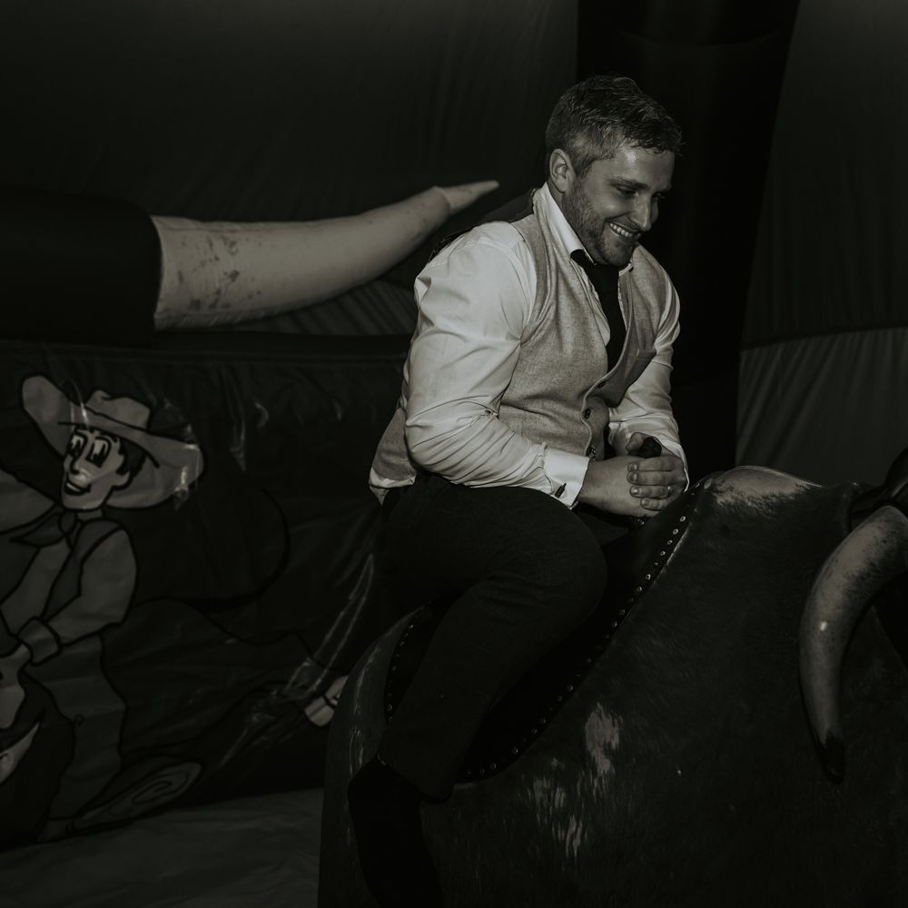 Groom in beige waistcoat on bucking rodeo bull at rustic barn wedding