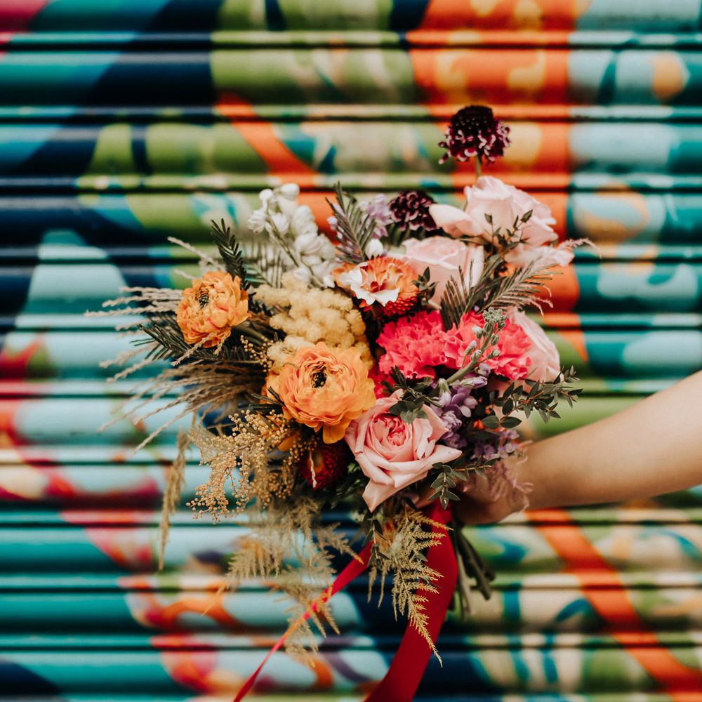 Bride holds colourful mixed bridal bouquet with pink roses tied with red ribbon infront of colourful background in streets of Liverpool