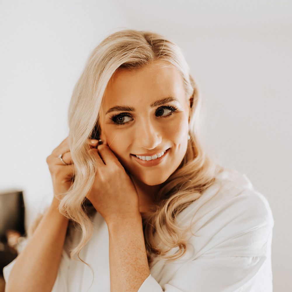 Bride with blonde curled hair and white bridal robe smiles as she puts in earrings before wedding