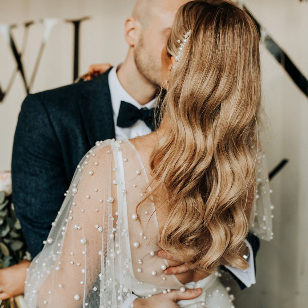 Bride with long curled hair and pearl hair clip wearing wedding dress with mesh pearl overlay kisses groom in navy tweed suit and bow tie