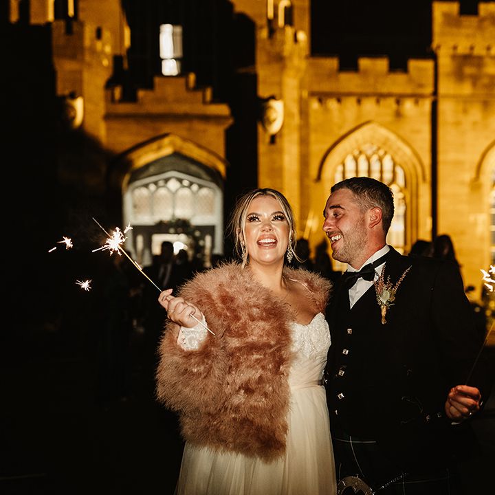 Bride looks up at fireworks and groom looks at her as they wave their own sparklers around 