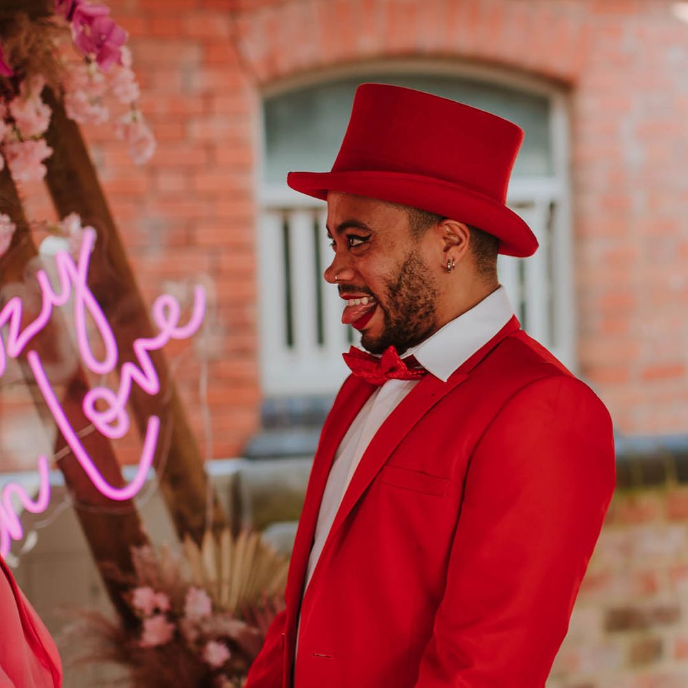 Stylish groom in a red suit and top hat wearing winged eyeliner and red lipstick makeup