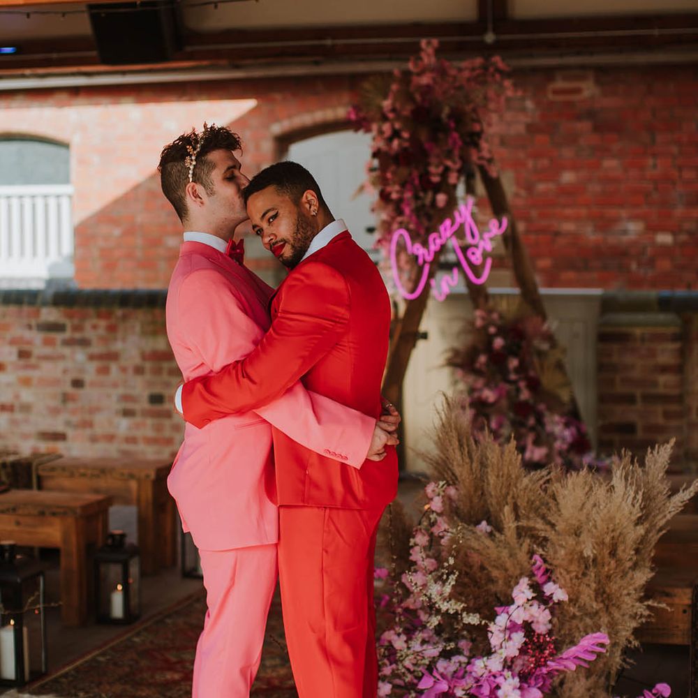 Groom in a pink suit, white boots and gold crown kissing his partners head in a red suit at Prestwold Hall Barns 