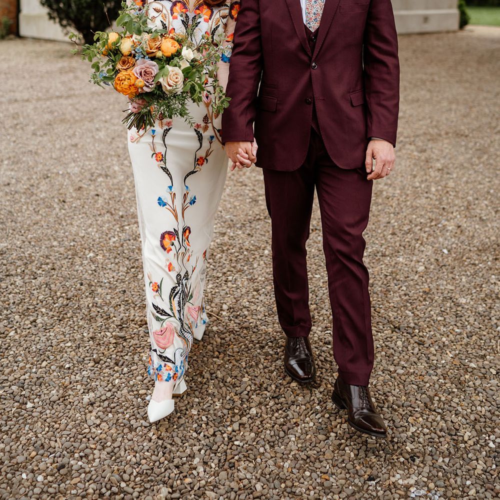 Groom in three piece burgundy suit with patterned tie with bride in white wedding dress with bright and colourful flowers 