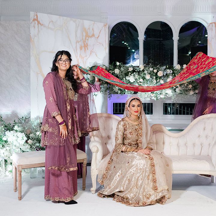 Bride sits on white sofa as wedding guests hold brightly coloured fabric above her head after Walima & Nikkah 