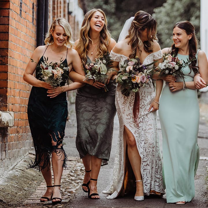 Bride in a boho lace wedding dress with bridesmaids in different shades of green dresses with small pink, red and purple flower bouquets