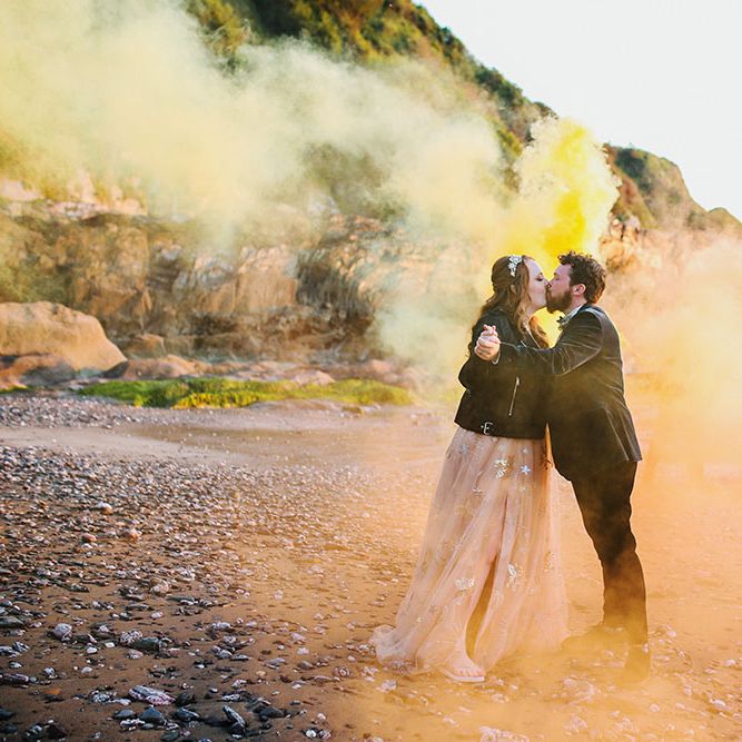 Bride wearing personalised leather jacket kisses groom whilst stood within orange and yellow smoke bombs along the beach