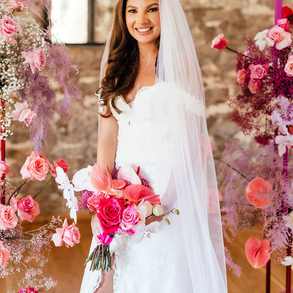 Bride stands between bright pink floral arch and wears lace sweetheart neckline wedding dress with floor length cathedral veil 