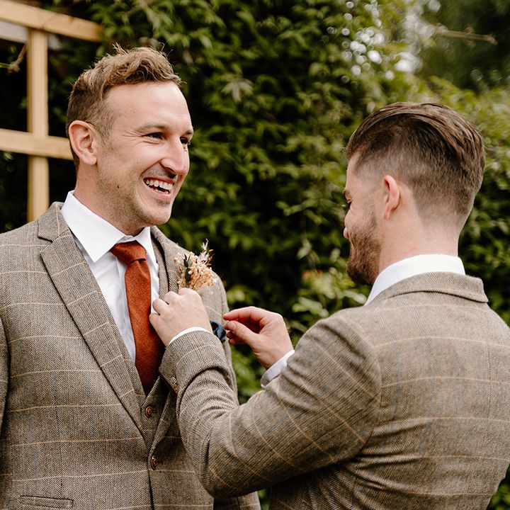 Groomsmen puts on the groom's dried flower buttonhole for him as they wear matching brown suits with orange ties