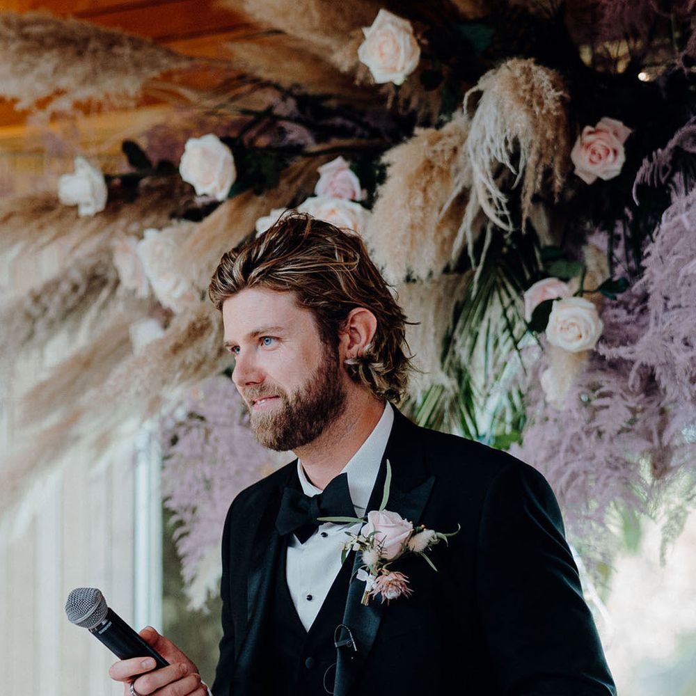 Groom in black tie with a pink rose buttonhole with a flower cloud in the background reads out his speech 