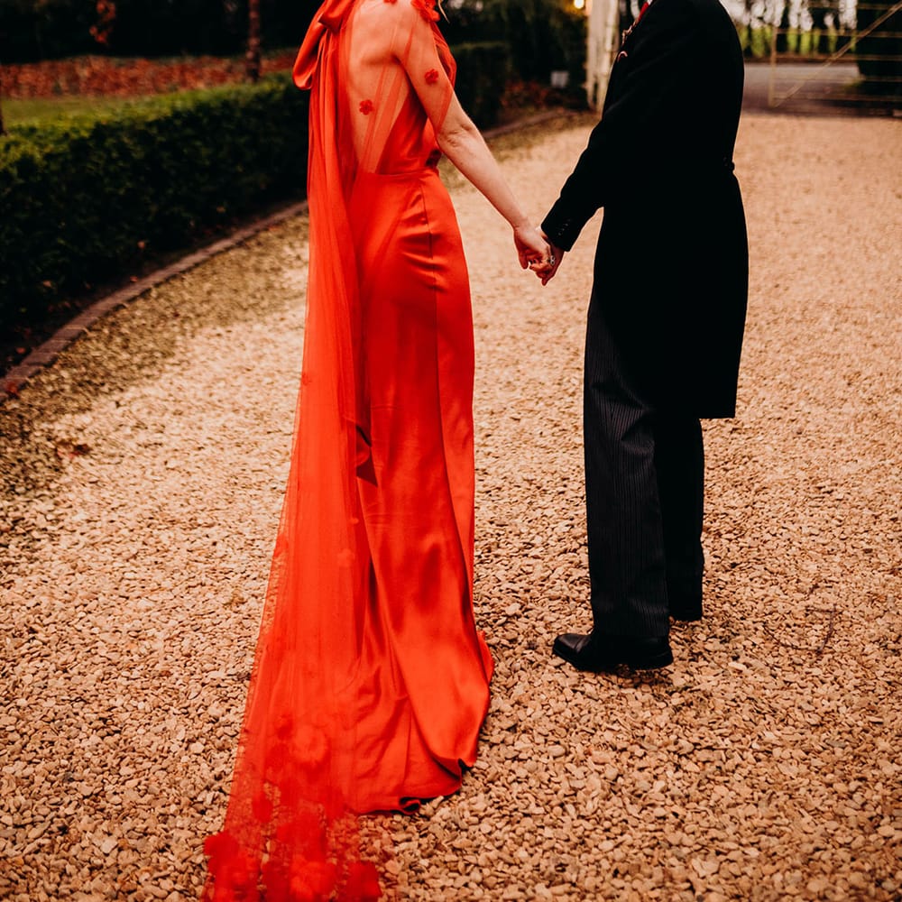 Bride in red satin wedding dress with cape and groom in three piece suit with red tie holding hands looking back at the camera