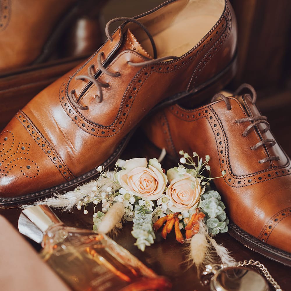Brown brogue wedding shoes beside pastel peach rose buttonhole and silver pocket watch 