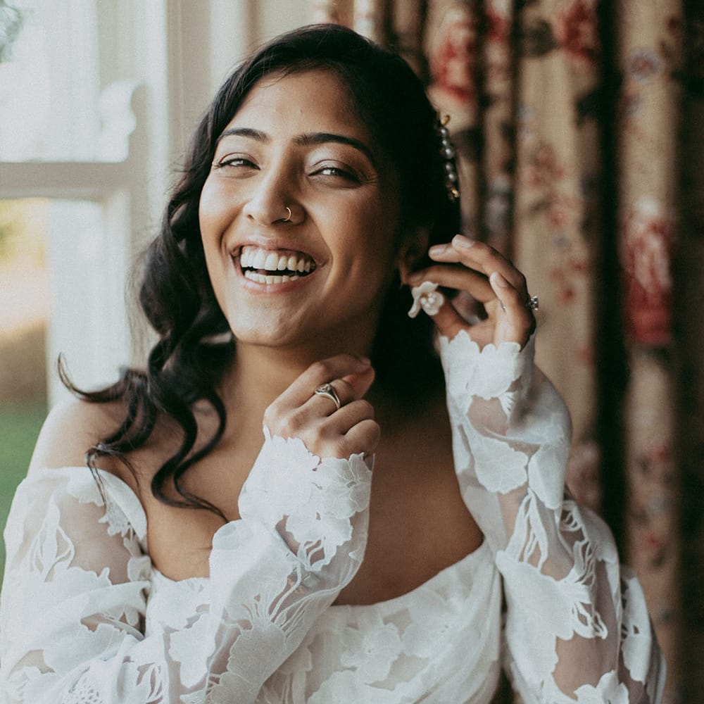 Bride in off shoulder long sleeve wedding dress with lace floral details adjusting white flower earrings 
