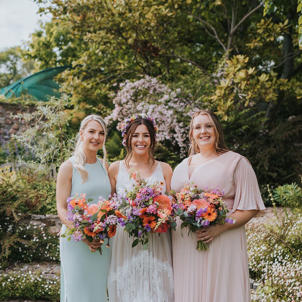 Bride stands besides her bridesmaids in pastel pink and mint green mismatched bridesmaid dresses holding colourful floral bouquets 
