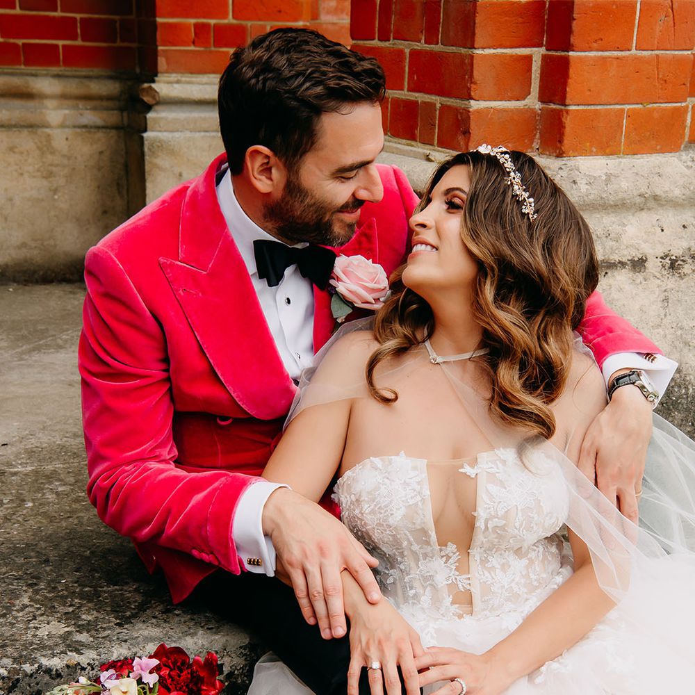Groom in bright pink velvet suit jacket and light pink rose buttonhole sitting on the stairs with the bride in a strapless wedding dress 